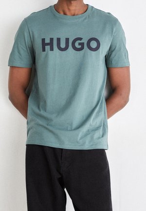 Camiseta de algodón teal de manga corta con un bold logo negro "HUGO" en el pecho, combinada con pantalones negros. Diseño simple y casual.