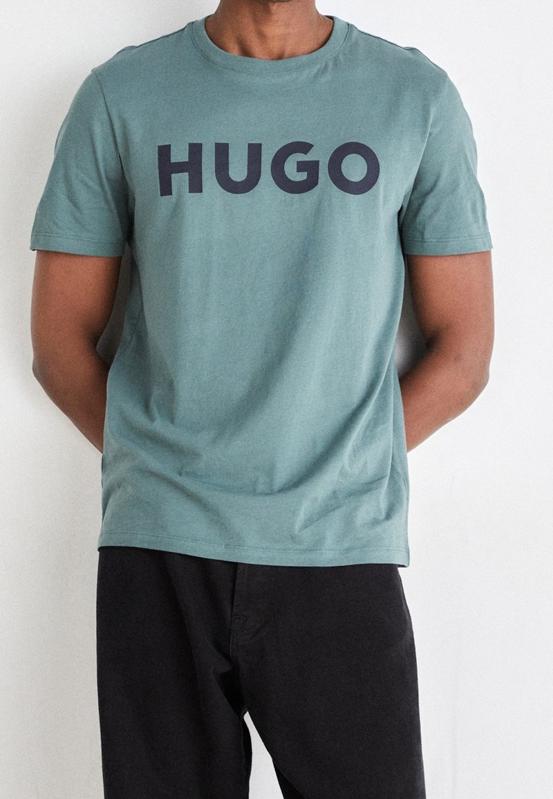 Tričko z teal bavlny s krátkým rukávem a výrazným černým logem "HUGO" přes hrudník, doplněné černými kalhotami. Jednoduchý, ležérní design.