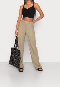Černý crop top s nařasením, béžové kalhoty s širokými nohavicemi a černá taška přes rameno s bílou linkovou kresbou. Celý outfit doplňují ploché sandály.