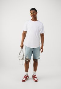 Hvit crew neck t-skjorte, lys blå slitte denimshorts, rød og hvit joggesko, holder en liten grå veske med stropp. Gullsolbriller.