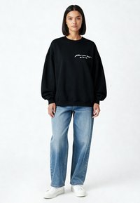 Vrouw staand, draagt een zwart sweatshirt met witte tekst, lichtblauwe spijkerbroek en witte sneakers, tegen een effen lichte achtergrond.