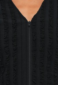 Nærbillede af et sort beklædningsstykke med lodrette flæser og en centreret sort lynlås over en tan hudbaggrund.