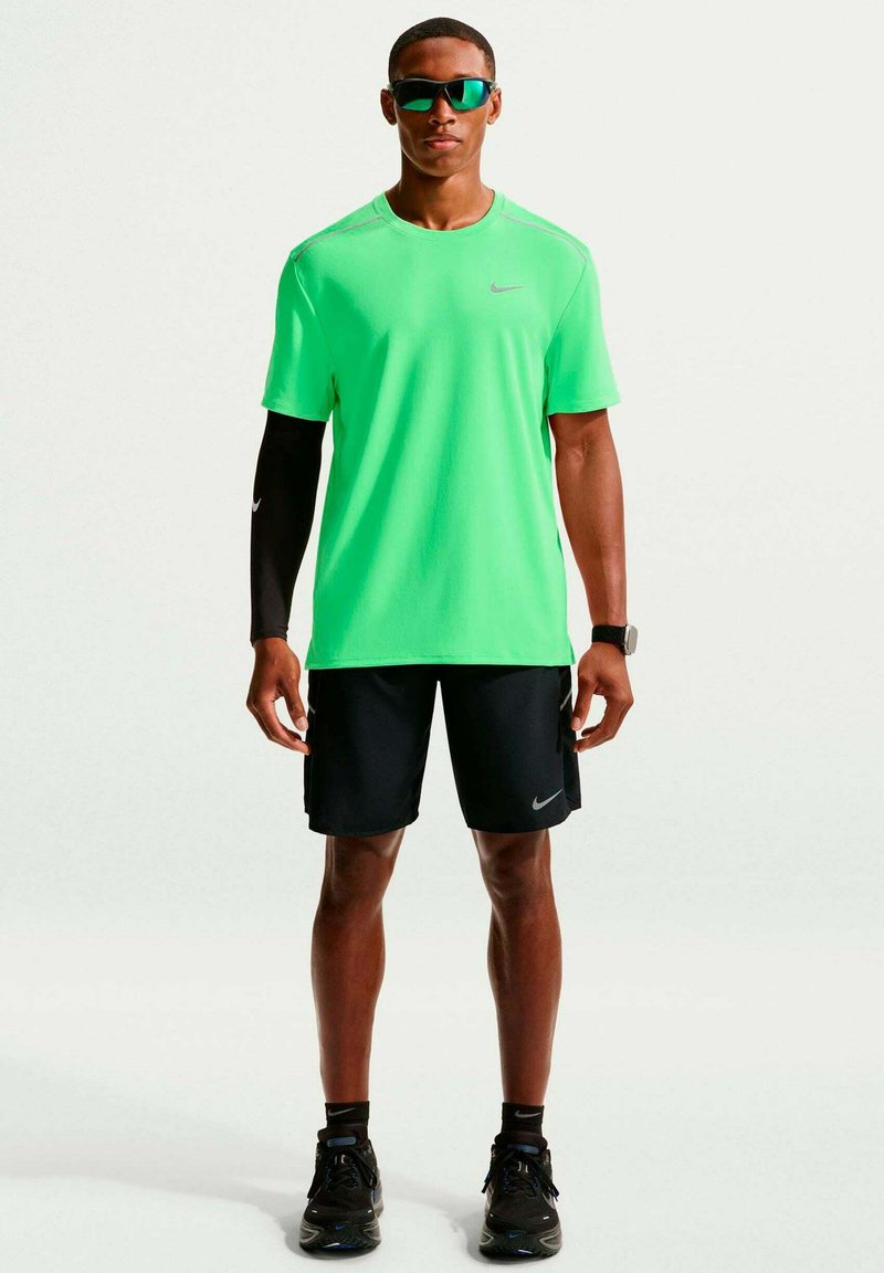 Man wearing neon green athletic shirt, black shorts, black running shoes, sunglasses, and a black arm sleeve on one arm standing facing forward.