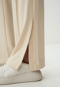 Pantalons beiges à jambe large en tissu doux avec une fente sur le côté. Associés à des baskets blanches texturées avec une semelle épaisse.