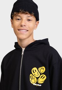Čierna mikina s kapucňou na zips s mäkkou textúrou a grafikou žltých usmievajúcich sa tvárí na prednej strane. Model nosí čiernu rebrovanú čiapku.
