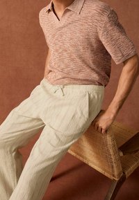 Homme portant un polo en maille à manches courtes texturé de couleur rouille et un pantalon à cordon blanc cassé, appuyé contre une chaise en bois tissé devant un fond marron.