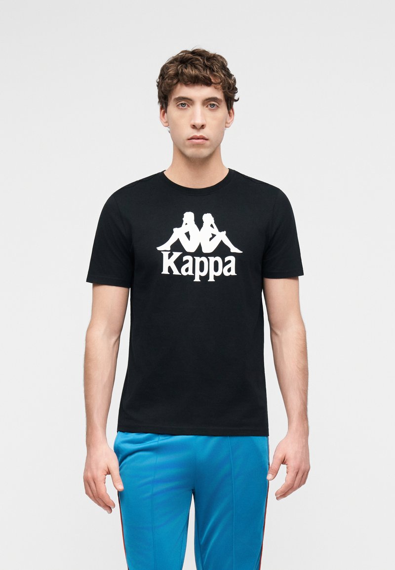 Νεαρός άνδρας που φοράει μαύρο μπλουζάκι Kappa με λευκό λογότυπο και μπλε παντελόνι, στέκεται μπροστά σε απλό ανοιχτό φόντο.