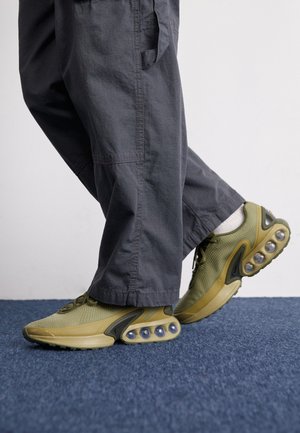 Personne portant un pantalon ample gris foncé et des baskets vert olive avec des semelles à coussin d’air visibles, debout sur un tapis bleu.