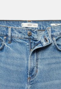 Jean en denim bleu avec une fermeture éclair et un bouton ouverts, avec des passants pour ceinture et des étiquettes de marque visibles à l'intérieur de la ceinture.