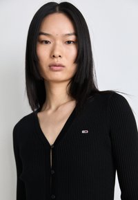 Melnas ribotas džemperis ar V veida kakla izgriezumu un piecām pogām priekšā, ar mazu krāsainu logo kreisajā pusē. Mīksta tekstūra un pieguļoša forma.