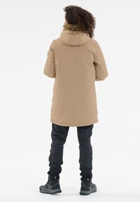 Beige parka dengan penutup kepala berbulu, memiliki potongan panjang, terbuat dari bahan tahan air, dan dipadupadankan dengan celana hitam serta sepatu boot hitam yang kokoh.