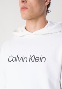 Hanorac alb dintr-un material moale, cu un buzunar mare tip kangaroo în față și un logo mare, negru "Calvin Klein" imprimat pe piept.