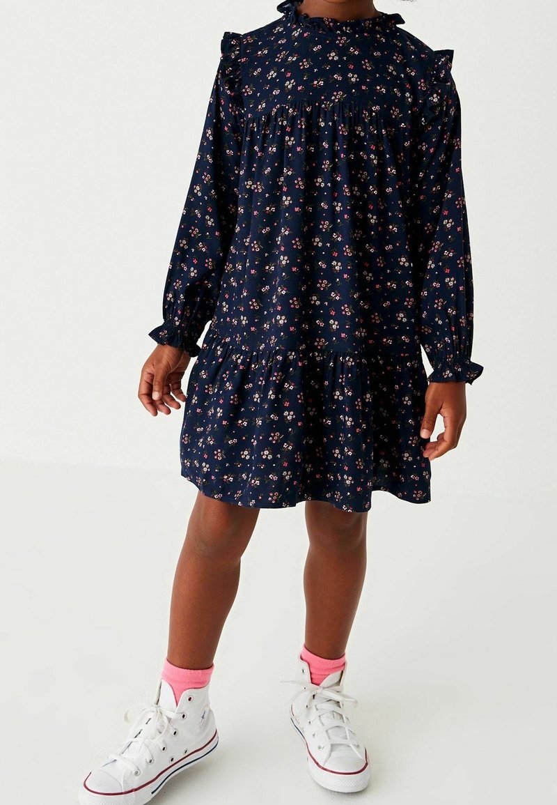 Enfant portant une robe longue manches longues bleu marine à motif floral avec volants, des chaussettes roses et des baskets montantes blanches, debout sur un fond uni.