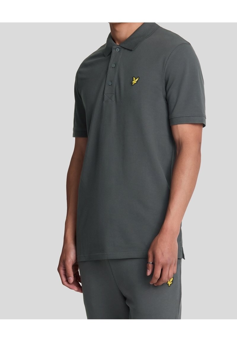 Homme portant un polo gris foncé à manches courtes et un pantalon assorti avec un logo oiseau jaune sur la poitrine et la cuisse, sur fond uni.