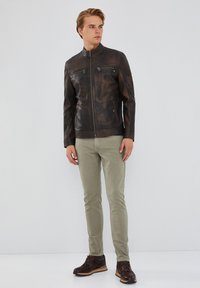 Bruine leren jas met een hoge kraag, twee met rits afsluitbare borstzakken en een gestructureerde afwerking. Gecombineerd met lichtgroene broek en donkere laarzen.