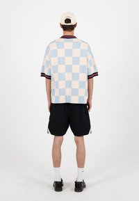 Mand iført en blå og beige ternet kortærmet skjorte, sorte shorts, hvide sokker, sorte sko og en beige kasket, stående med ryggen mod fronten.