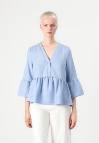 Lyseblå bluse i linblanding med V-hals og fem knapper. Har ermelengde med volanger og en samlet peplumliv.