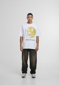 Hvid oversized t-shirt med et gult smiley ansigt design og teksten "Produkt af Internettet." Kombineret med mørke, posede jeans og sneakers.