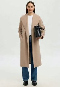 Manteau long beige avec un col châle, associé à une chemise blanche et un jean bleu. Le mannequin tient un sac à main noir structuré. Des chaussures noires complètent le look.