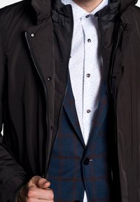 Dark, waterproof outer jacket with a zip, layered over a blue checkered blazer and white shirt with small blue dots and brown buttons.