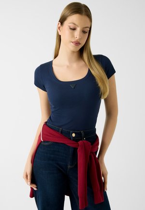 Jeune femme portant un haut ajusté bleu marine à manches courtes et un jean foncé, avec un pull rouge noué autour de la taille, regardant vers le bas.