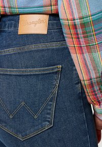 Mörkblå denimjeans med en framträdande Wrangler-läderlogotyp, gula sömmar och en distinkt triangulär bakfickedesign.