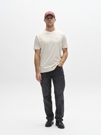 Beige T-shirt met korte mouwen en een subtiel merklogo, gecombineerd met donkergrijze jeans en zwarte schoenen. Model draagt een bordeauxrode pet. Eenvoudige achtergrond.