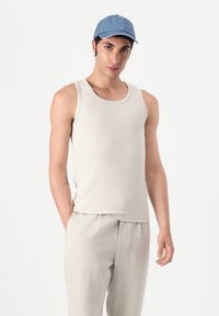 Beige sleeveless tank top with a soft texture, paired with light beige pants. A blue cap sits atop dark hair. Simple design with no patterns.