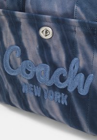 Modra tkaninska torba z izbočeno, vezeno oznako "Coach New York". Vključuje kovinsko zapiranje na pritisni gumb in teksturirano površino.