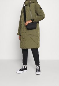 Parka acolchada verde oliva con capucha, bolsillos frontales con cremallera y bolso de hombro negro. Pantalones negros y zapatillas negras de caña alta completan el conjunto.