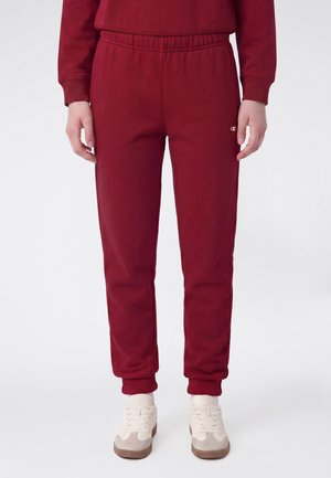 Persona che indossa pantaloni da jogging bordeaux e felpa abbinata con sneakers bianche, in piedi davanti a uno sfondo neutro.
