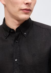 Ανδρικός κορμός που φοράει μαύρο πουκάμισο με κουμπιά και μικρό κεντημένο λογότυπο στο στήθος, με το γιακά κουμπωμένο.