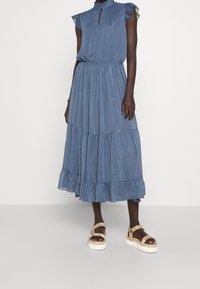 Blauwe midi-jurk met hoge hals, gerimpelde mouwen en gelaagde rok. Gemaakt van textiel met een structuur en metallic strepen; gecombineerd met tan platform sandalen.