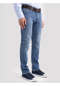 Mees, kandes helesiniseid teksapükse, helesinist särki, pruuni vööd ja tumelikke siniseid tossusid, seisab tavalise tausta ees.