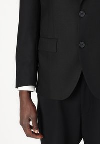 Veste de costume noire en tissu texturé à simple boutonnage, avec une poche poitrine, deux poches à rabat, et boutons noirs. Manche de chemise blanche visible.