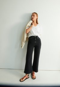 Zwarte flared jeans, witte V-hals T-shirt, crèmekleurige jas. Sandalen met rode banden. Model staat tegen een lichte muur.
