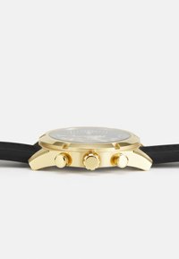 Goldfarbener Chronograph mit einem schwarzen Silikonarmband, strukturiertem Zifferblatt, drei Drückern und einem flachen Mineralglas-Uhrenglas.