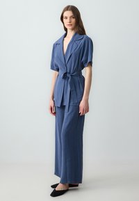 Completo due pezzi azzurro chiaro composto da un blazer a maniche corte con cintura e pantaloni a gamba larga realizzati in un tessuto testurizzato. Scarpe nere senza lacci.