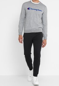 Felpa in pile grigia con logo "Champion" blu, collo a giro, polsini a costine, abbinata a pantaloni jogger neri e sneakers bianche.