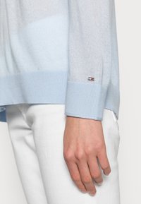 Helles blaues Strickpullover mit gerippten Bündchen. Verfügt über ein kleines Logo-Detail am Handgelenk. Kombiniert mit weißen Hosen, mit Fokus auf dem Ärmel und der Hand.