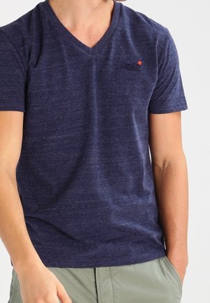Mand iført en mørkeblå V-hals T-shirt og lysegrønne bukser, med den ene hånd i lommen mod en ensfarvet baggrund.