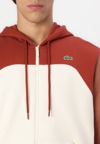 Hettejakke med glidelås som har et delt design, med en rustfarget overdel og en hvit underdel, akkompagnert av en liten grønn logo.