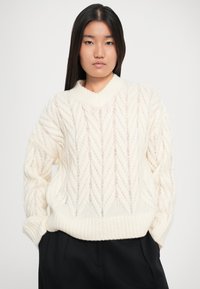 Pull en tricot crème avec un motif en chevrons, col et poignets côtelés, coupe oversize et design en maille ouverte texturée.