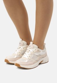 Zapatillas deportivas beige con paneles de malla y cuero, punta redonda, cordones blancos y una suela de goma texturizada. Presenta un logo rosa claro en la lengüeta.