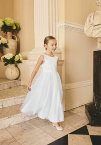 Junges Mädchen in weißem Kleid und Schuhen, das eine Marmor-Treppe in einem eleganten Raum mit klassischen Statuen und Blumenvasen hinabsteigt.