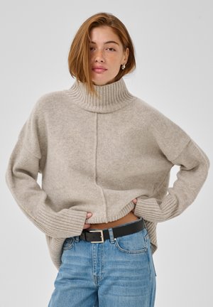 Jeune femme aux cheveux roux portant un pull à col roulé beige et un jean bleu avec une ceinture noire sur un fond clair uni.