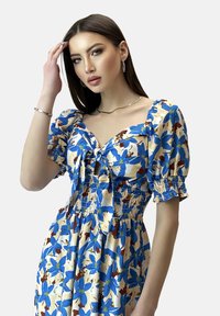 Blumenmusterkleid mit Puffärmeln, das blaue und cremefarbene Muster auf einem hellen Hintergrund zeigt. Rüschenoberteil mit einer Bindefront.