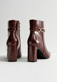 Botas de tobillo de cuero burdeos con acabado brillante, tacones altos de bloque y detalle de hebilla dorada en el costado. Textura suave, cierre con cremallera.