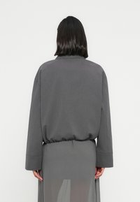 Άτ ομο με σκούρα μαλλιά μέχρι τους ώμους, φοράει φαρδιά γκρι ζακέτα και διαφανή γκρι φούστα, στέκεται μπροστά σε λευκό, απλό φόντο.