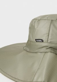 Chapeau seau vert olive fabriqué en tissu r�ésistant à l'eau, avec un large bord et un petit logo noir "RAINS" sur le côté. Finition texturée.
